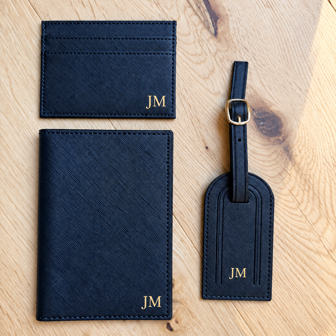 A dark navy blue leather luggage set with gold monogram initials 'JM' on a light wood background. The set includes a passport cover, a card holder, and a luggage tag with a gold buckle.