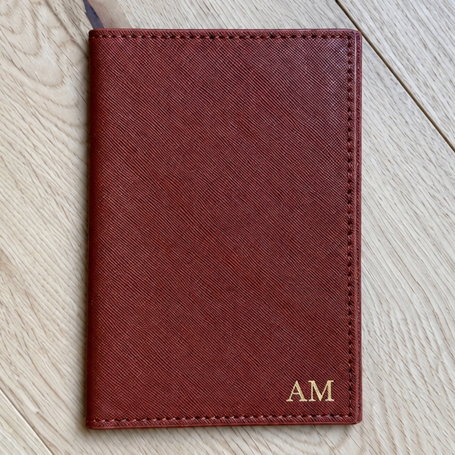 A reddish-brown leather passport cover with gold 'AM' initials, laid flat on a light wood floor.