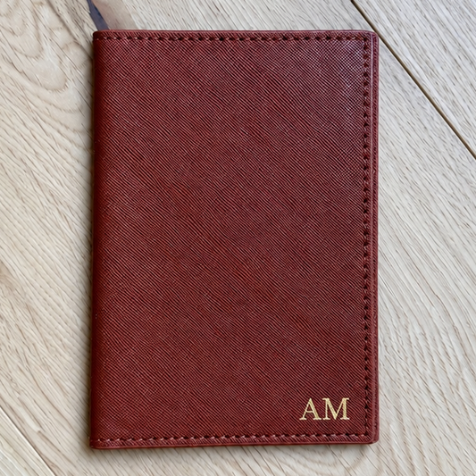 A reddish-brown leather passport cover with gold 'AM' initials, laid flat on a light wood floor.
