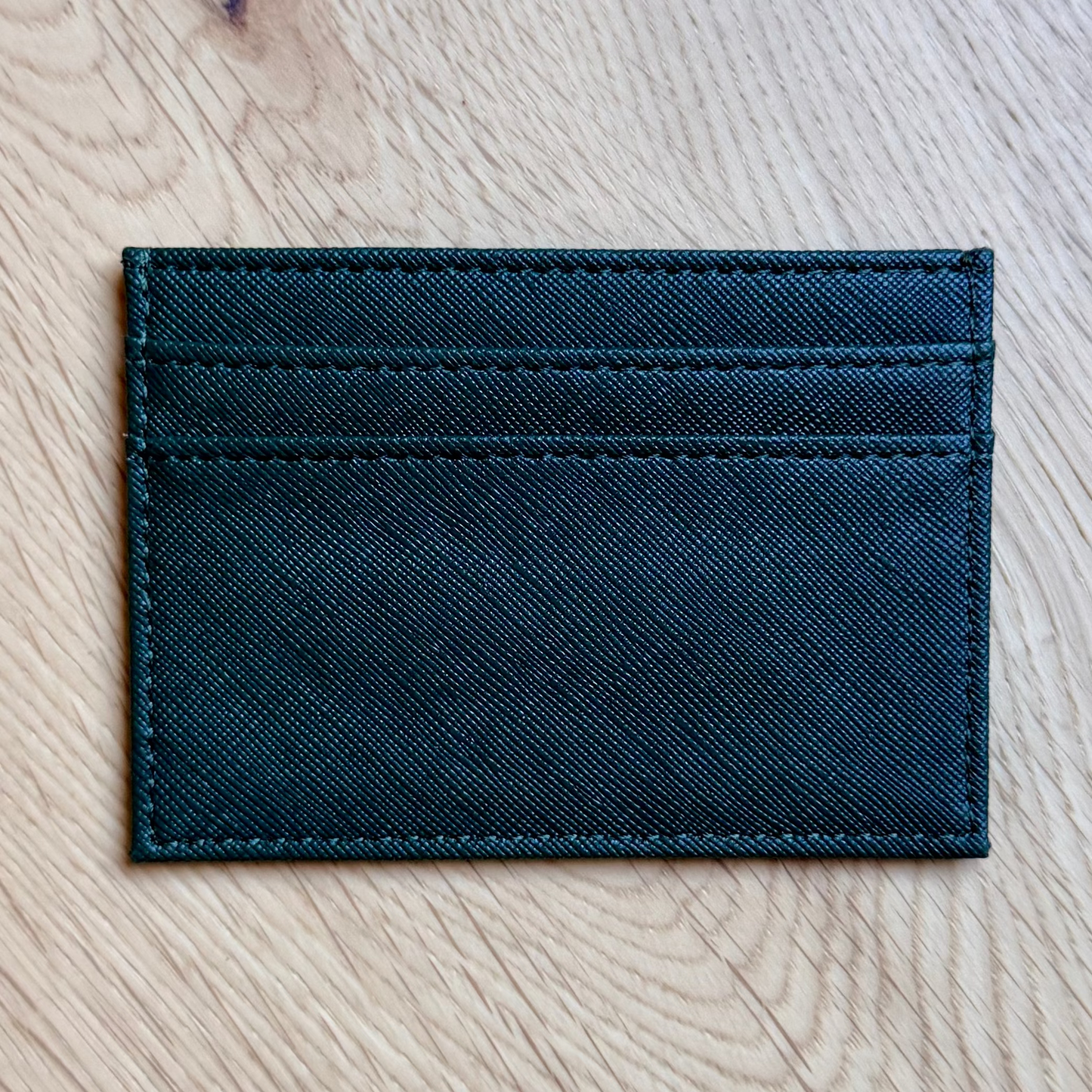A slim, dark teal leather card holder wallet with stitched details, placed on a light-colored wooden floor.