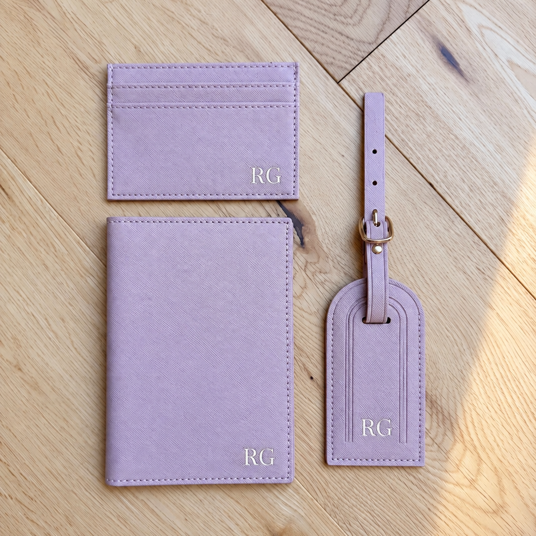 A coordinated set of light lavender leather luggage accessories on a light wood floor: a passport holder, a card holder, and a luggage tag with a strap and gold-tone hardware. Each item is embossed with the initials 'RG'.